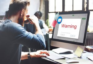 A man drinks a cup of coffee while looking at his work computer with a big warning box on it.