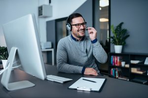 A man on a headset in front of a computer smiles as he speaks with a client.