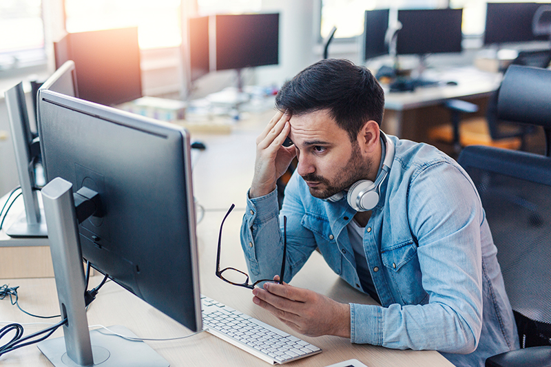 A man looks frustrated with his hand on his forehead, looking at a computer.