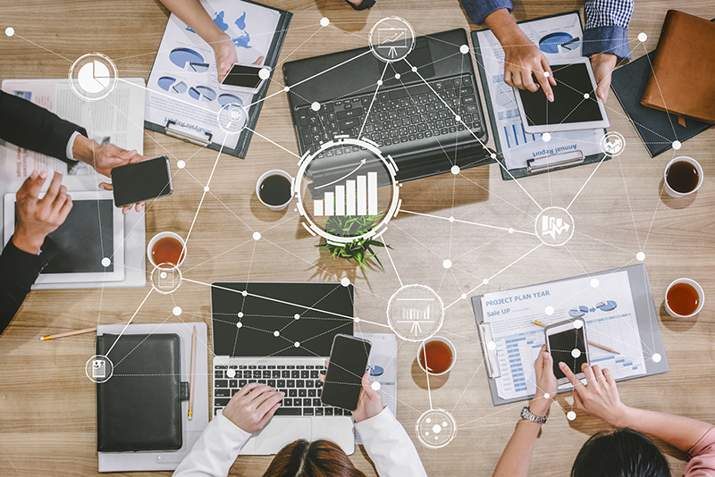 Multiple people gathered around a work table with reports, laptops, phones, and tablets and business growth icons overlaid on top.