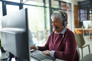 An older woman smiles while wearing a headset and working on a computer.