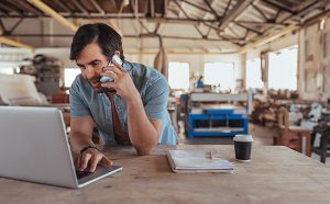 A man talking on a phone and working on his lap at a woodworking shop.