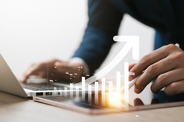 A person on a laptop with a graphic of a line chart over their hands and an upward arrow.