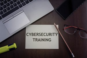 A desk with a laptop, pencil, glasses, and a note that reads ‘Cybersecurity Training.’