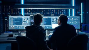 A view from behind of two IT professionals working at a set of three computer monitors in a network room